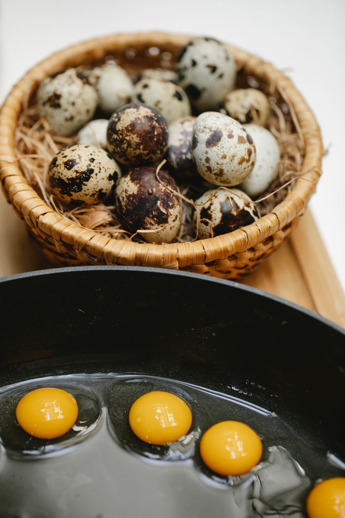 From above of many small eggs frying in black pan on wooden surface near wicker bowl with heap of quail eggs